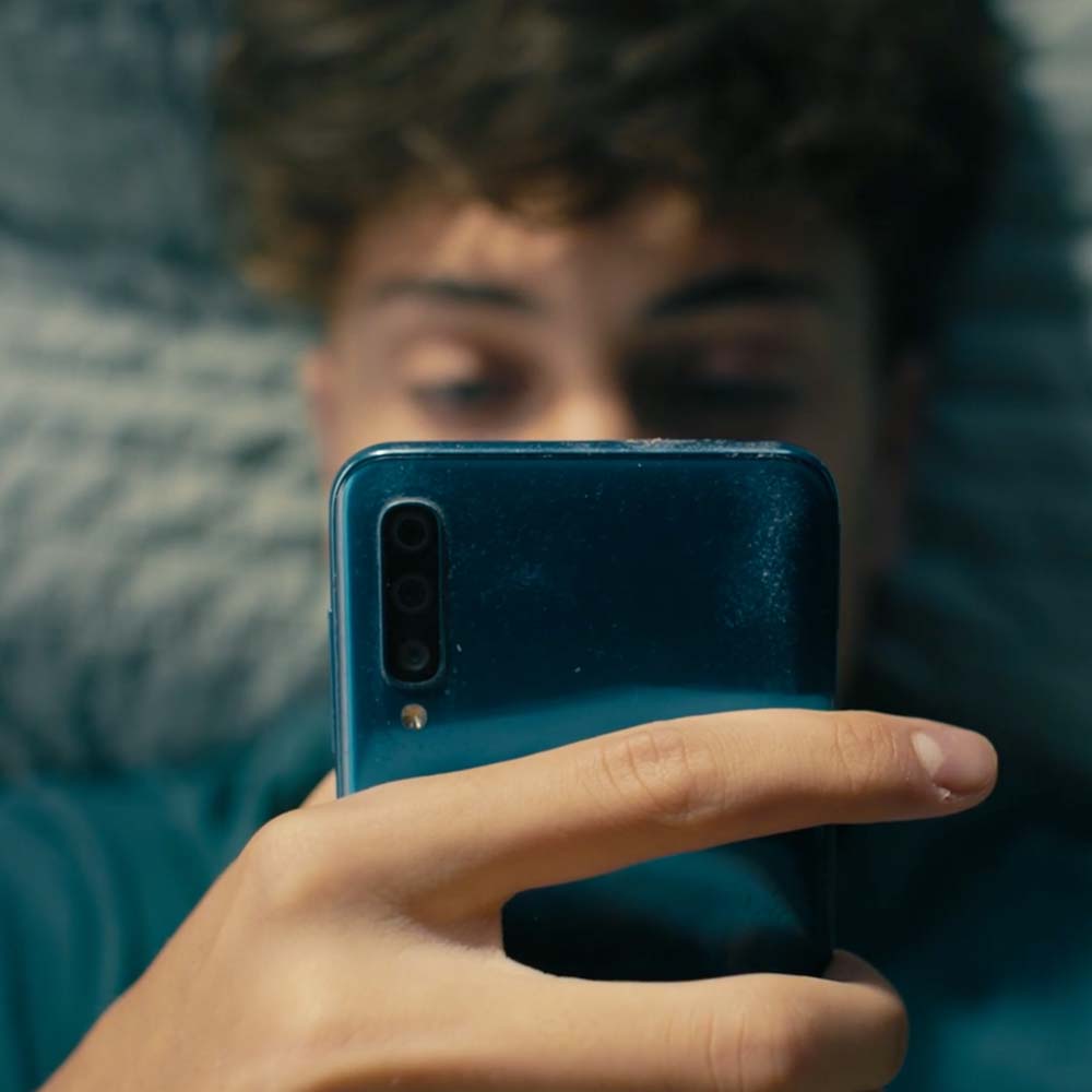 Ung mand ligger ned og kigger koncentreret på en blå smartphone, mens han doomscroller på sin telefon.