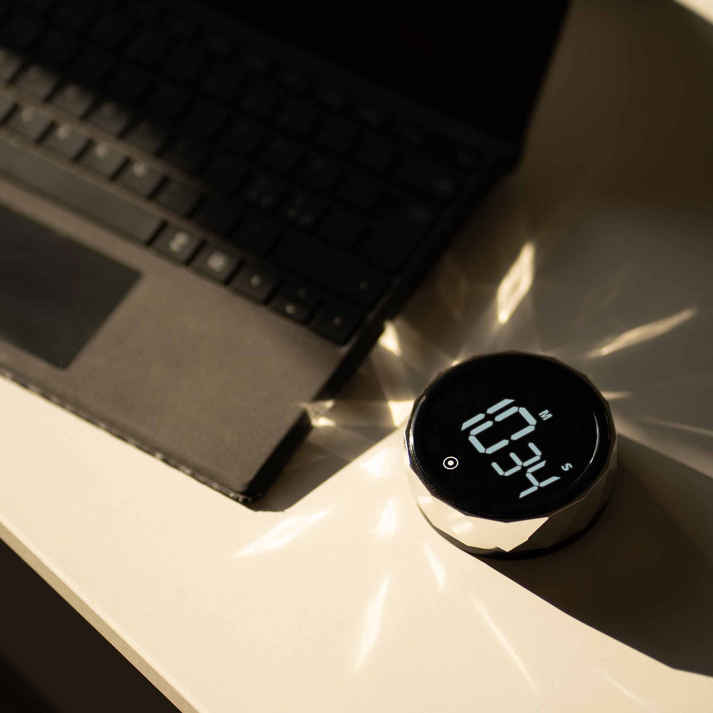 Futuristisk digitalt ur med facetteret sølvramme, placeret på et hvidt skrivebord ved siden af en sort bærbar computer, med sollys, der reflekteres dramatisk fra urets overflade.