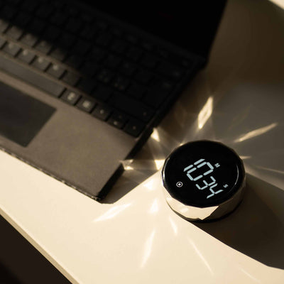 Futuristisk digitalt ur med facetteret sølvramme, placeret på et hvidt skrivebord ved siden af en sort bærbar computer, med sollys, der reflekteres dramatisk fra urets overflade.