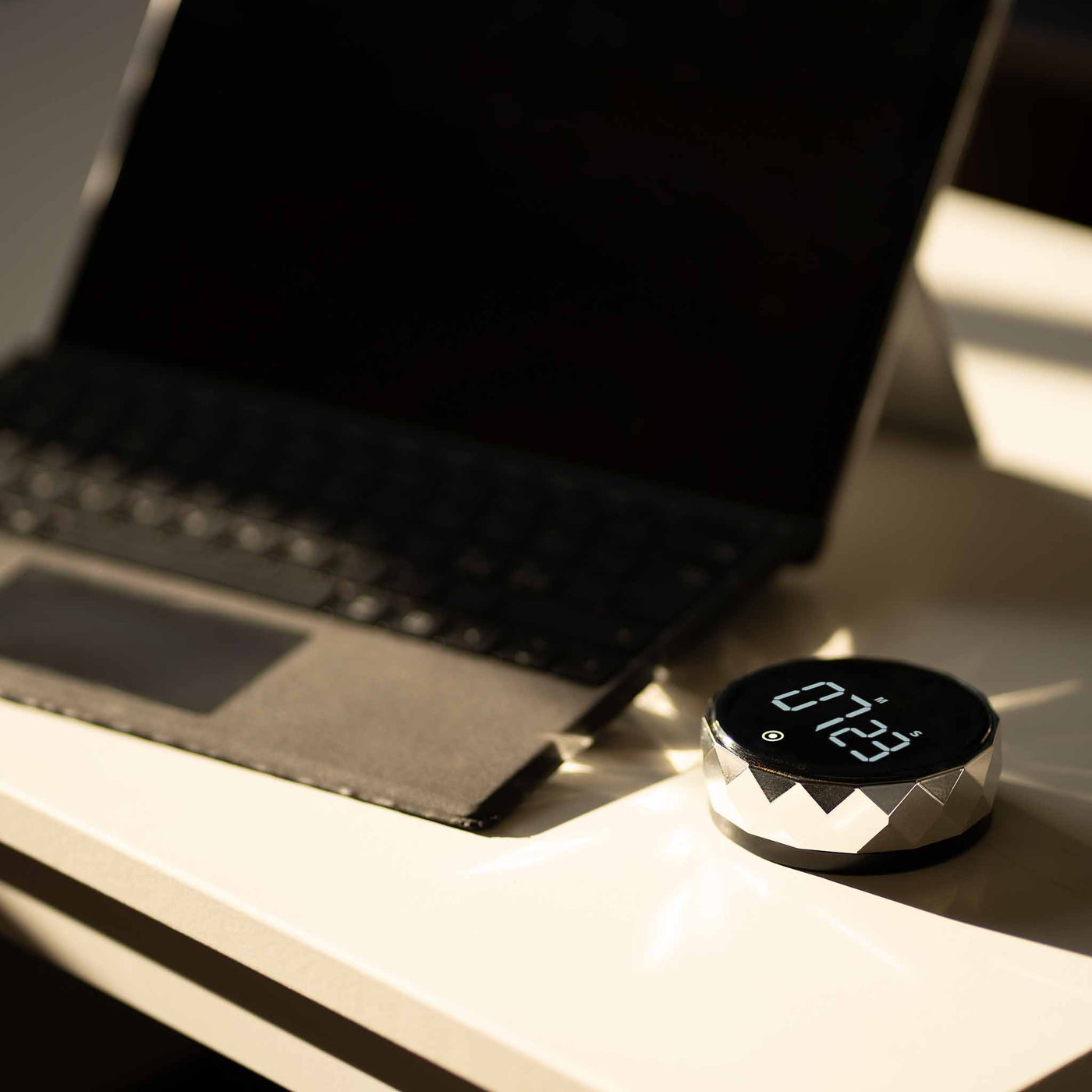 Futuristisk digitalt ur med facetteret sølvramme, placeret på et hvidt skrivebord ved siden af en åben bærbar computer, skaber et moderne og sofistikeret arbejdsmiljø.