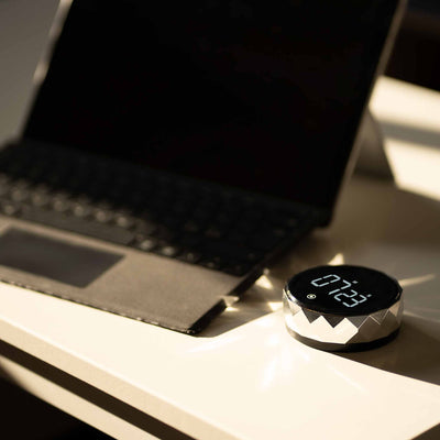 Futuristisk digitalt ur med facetteret sølvramme, placeret på et hvidt skrivebord ved siden af en åben bærbar computer, skaber et moderne og sofistikeret arbejdsmiljø.
